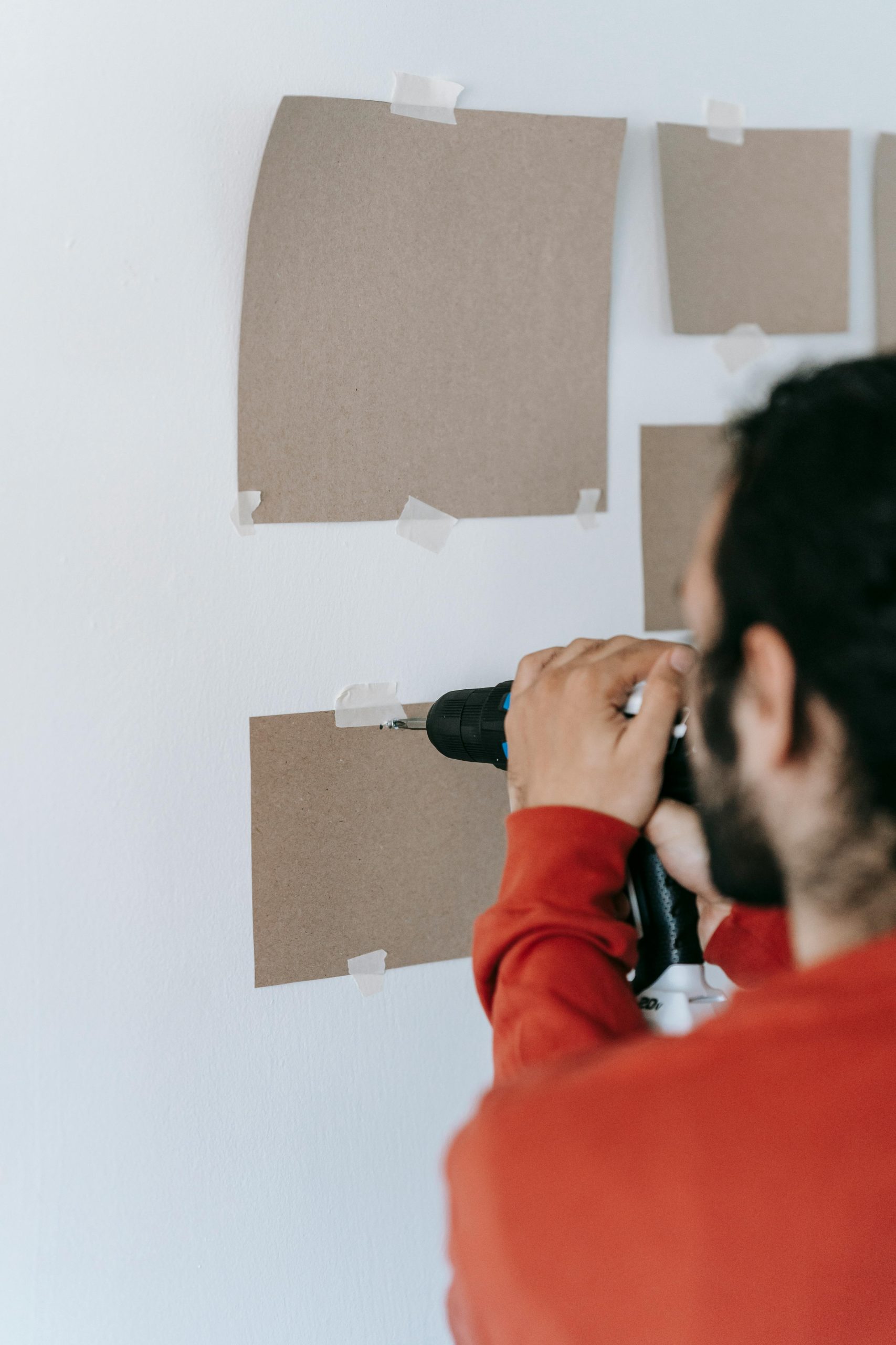 Vue arrière d'un homme barbu méconnaissable utilisant un tournevis électrique pour visser dans un mur avec des plaques de carton dans une maison.
