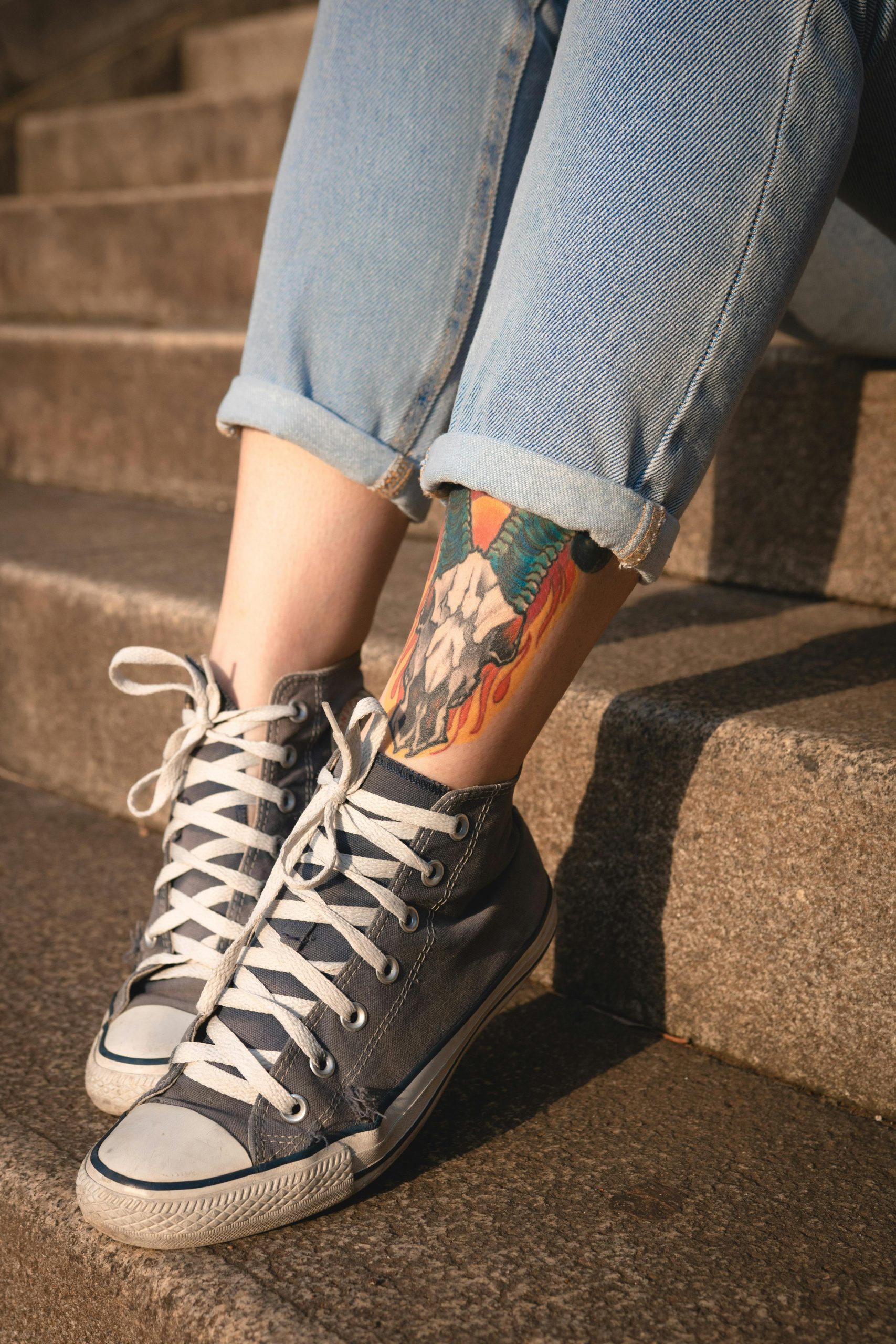 Close-up des jambes d'une femme en jeans et baskets, mettant en valeur un tatouage coloré sur la jambe dans un cadre urbain.