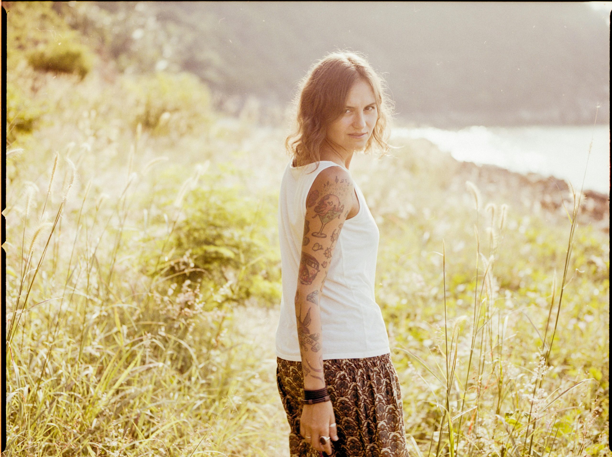 Une femme tatouée en débardeur blanc se tient dans un champ d'herbe en regardant le coucher de soleil.