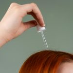 Gros plan d'une main appliquant du sérum capillaire sur des cheveux roux avec un compte-gouttes.