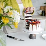 Crop unrecognizable female confectioner in casual clothes and apron serving yummy homemade cake with white cream and assorted fresh berries standing in light kitchen decorated with flowers vase and plant --- Pâtissière méconnaissable en tenue décontractée et tablier servant un délicieux gâteau maison avec crème blanche et baies fraîches assorties debout dans une cuisine lumineuse décorée d'un vase de fleurs et d'une plante