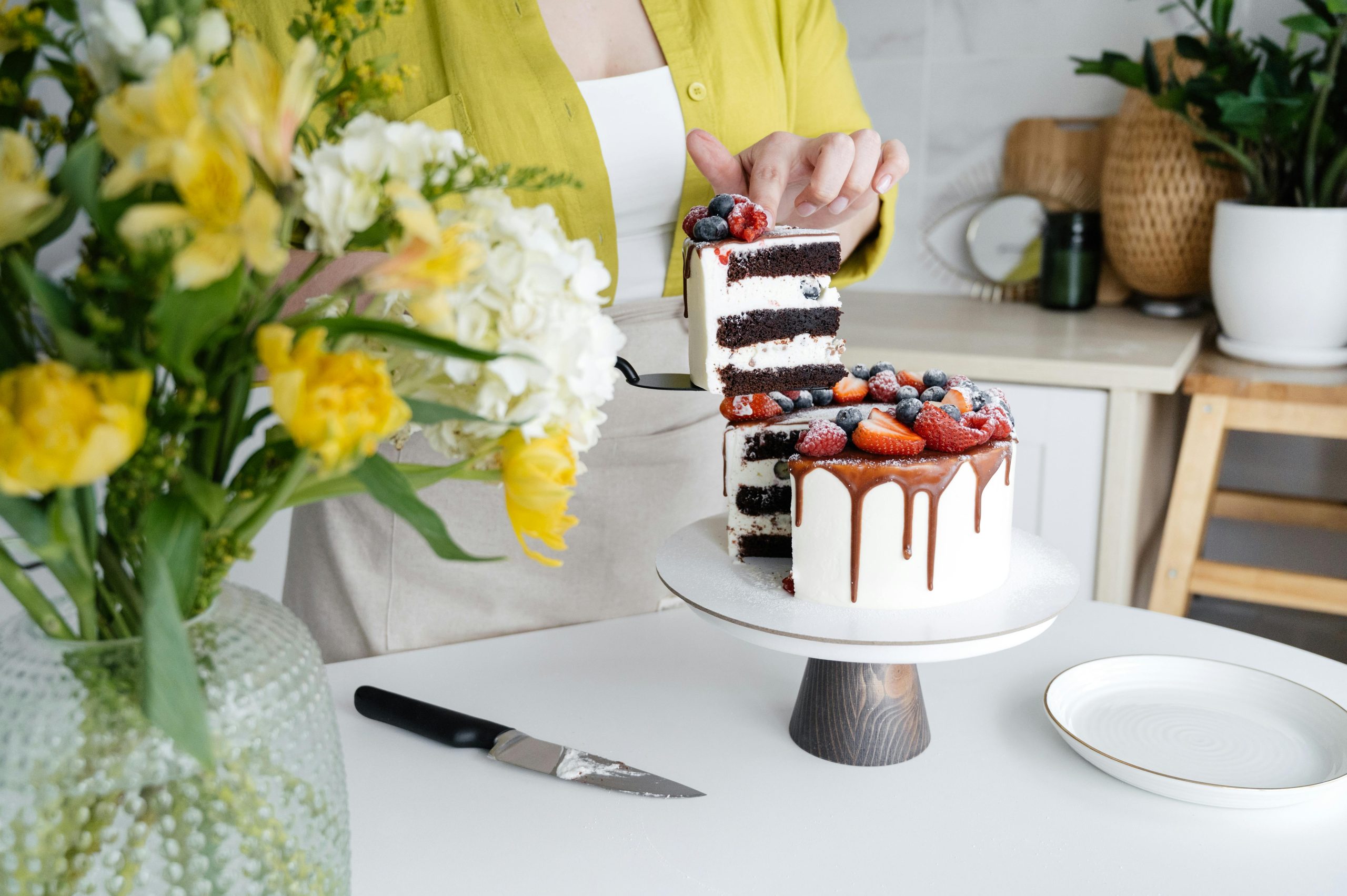 Crop unrecognizable female confectioner in casual clothes and apron serving yummy homemade cake with white cream and assorted fresh berries standing in light kitchen decorated with flowers vase and plant --- Pâtissière méconnaissable en tenue décontractée et tablier servant un délicieux gâteau maison avec crème blanche et baies fraîches assorties debout dans une cuisine lumineuse décorée d'un vase de fleurs et d'une plante