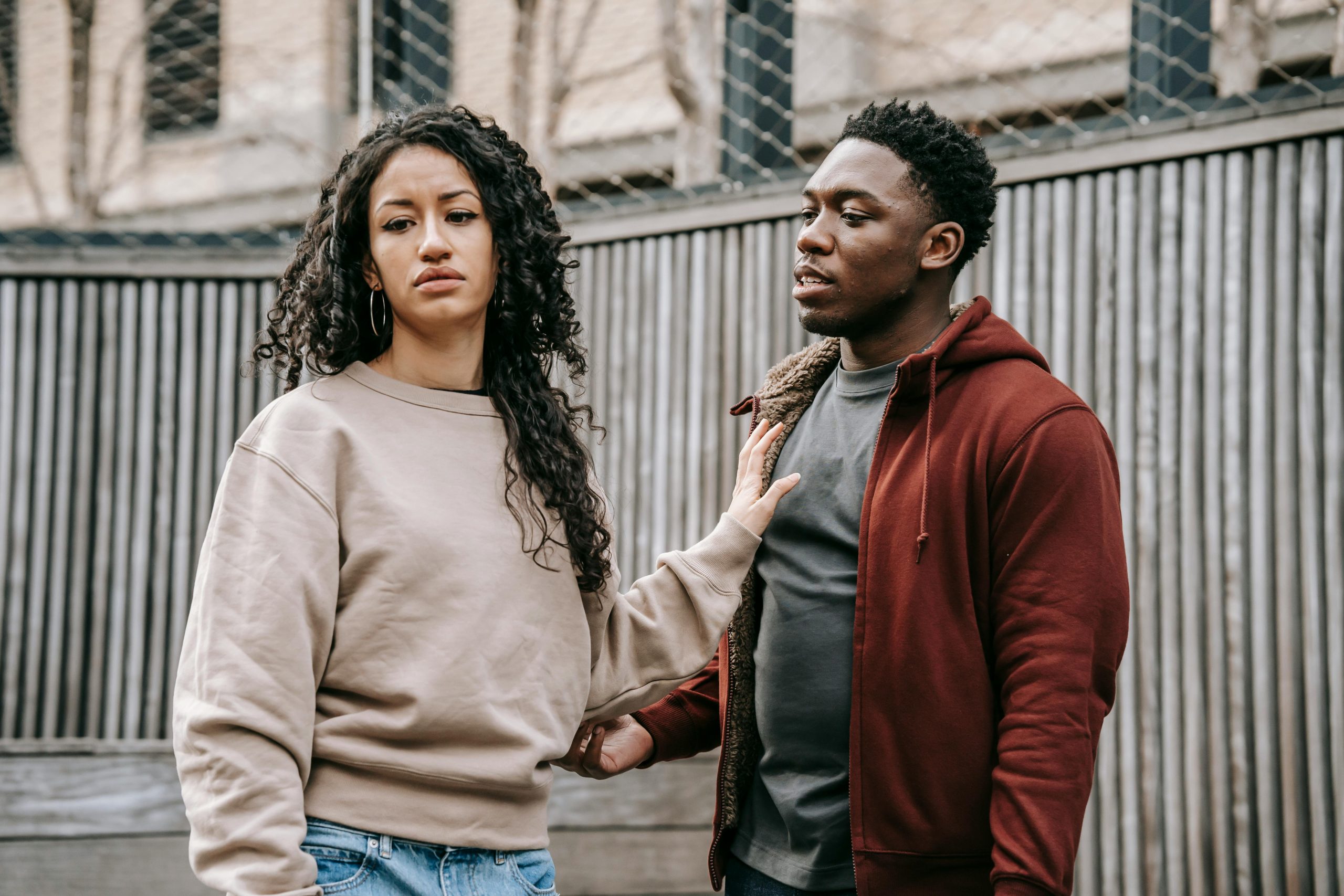 Sad ethnic girlfriend with curly hair rejecting annoyed African American boyfriend while arguing on street near wooden fence during breakup Petite amie ethnique triste aux cheveux bouclés rejetant un petit ami afro-américain ennuyé en se disputant dans la rue près d'une clôture en bois pendant une rupture