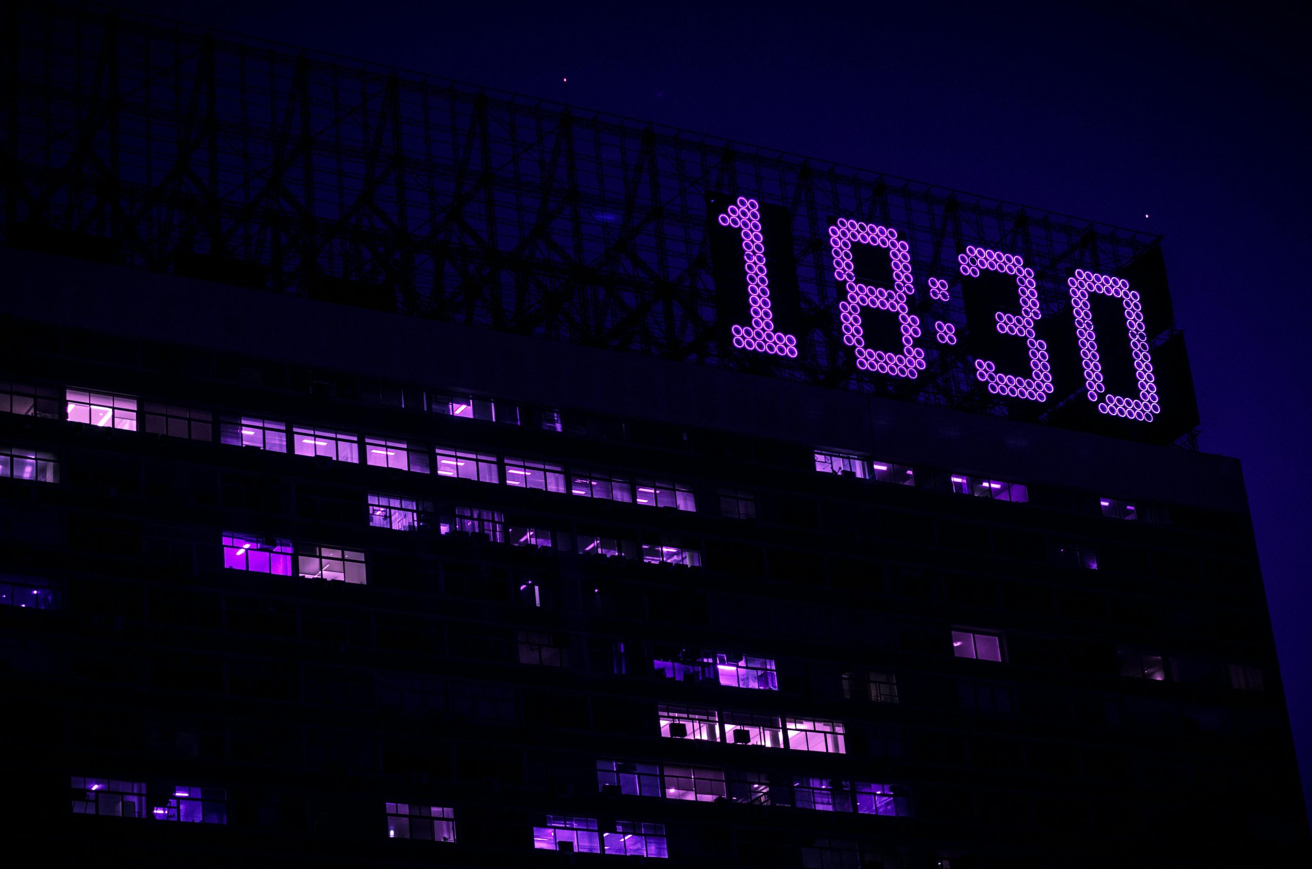 A modern building is illuminated at night with a prominent digital clock showing 18:30. Un bâtiment moderne illuminé la nuit avec une horloge numérique bien en vue affichant 18:30.