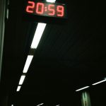 A brightly lit digital clock displaying 20:59 in a dark subway station setting. Une horloge numérique brillamment éclairée affichant 20:59 dans un environnement de station de métro sombre.