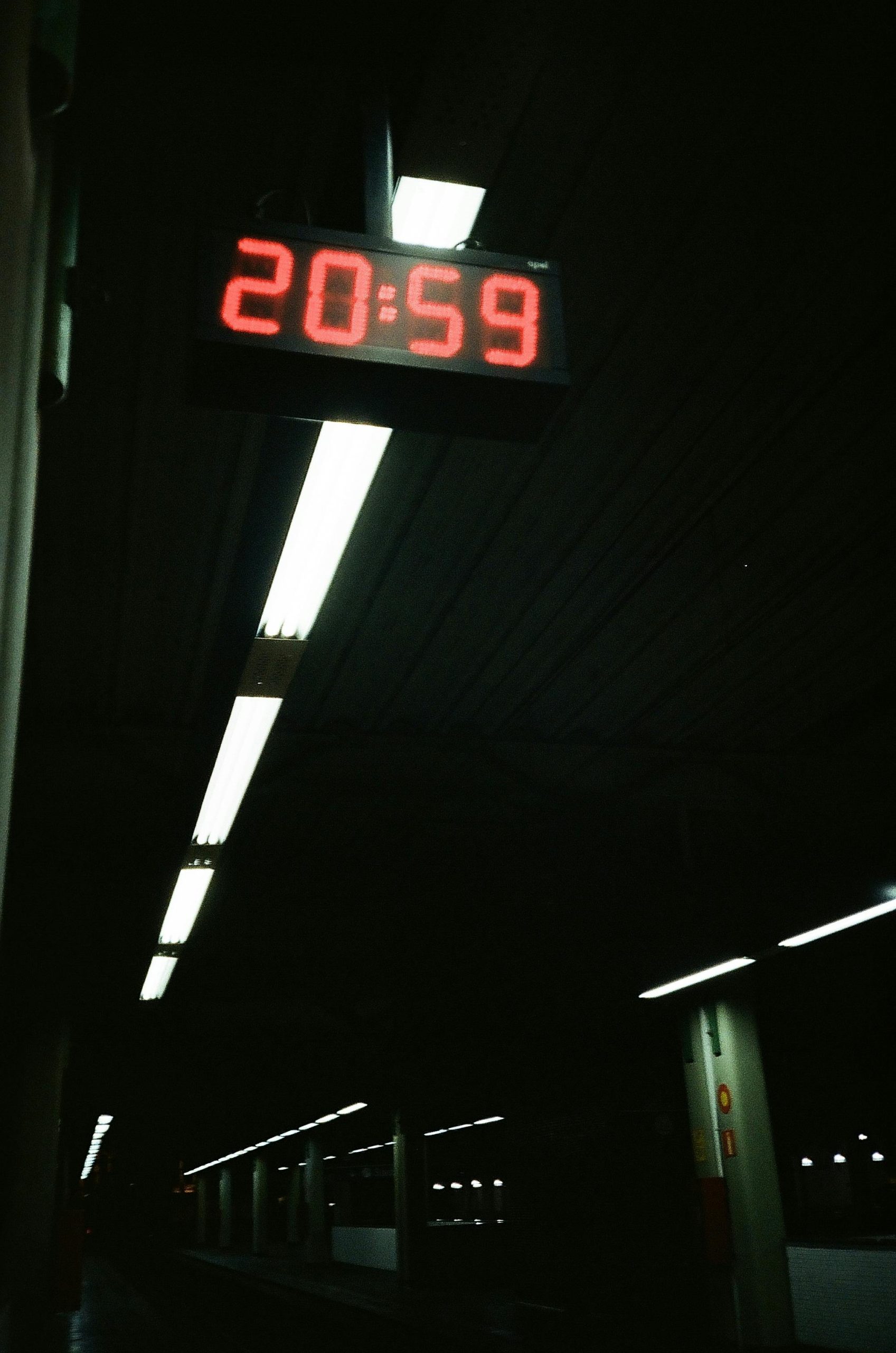 A brightly lit digital clock displaying 20:59 in a dark subway station setting. Une horloge numérique brillamment éclairée affichant 20:59 dans un environnement de station de métro sombre.