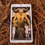 The Devil tarot card placed on dried pine needles, showing intricate artwork. La carte de tarot Le Diable placée sur des aiguilles de pin desséchées, montrant un artwork complexe.
