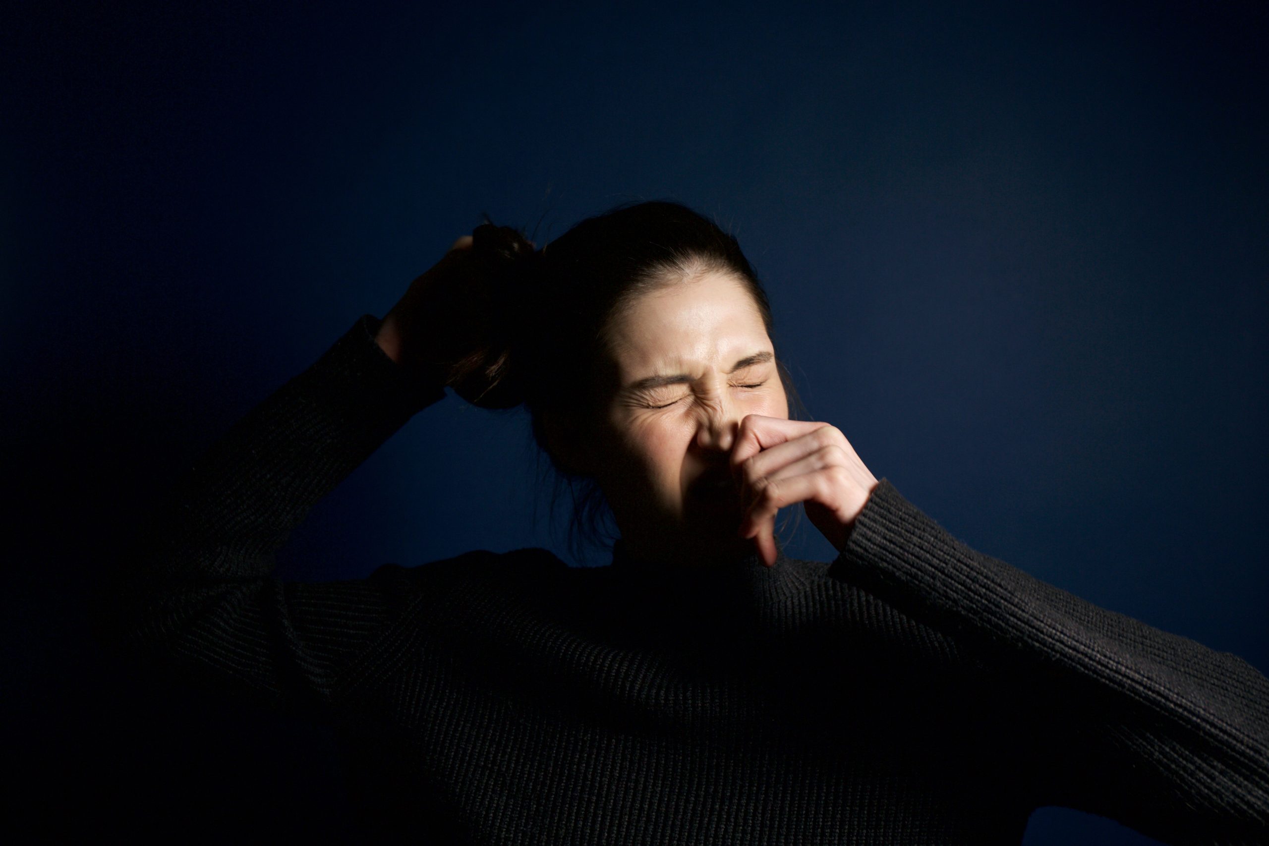 A woman in a dark sweater sneezes against a solid blue background, capturing a moment of candid expression. Une femme en pull foncé éternue sur un fond bleu uni, capturant un moment d'expression candide.