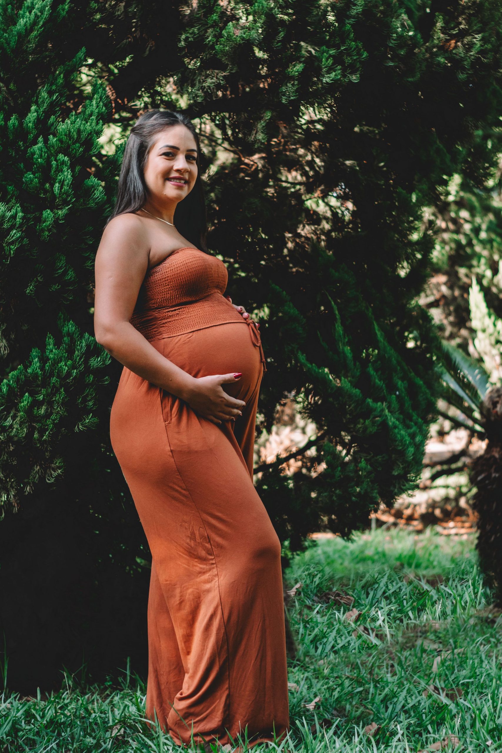 A pregnant woman standing and smiling in a lush outdoor setting, captured in a warm portrait. Une femme enceinte debout et souriante dans un cadre extérieur luxuriant, capturée dans un portrait chaleureux.