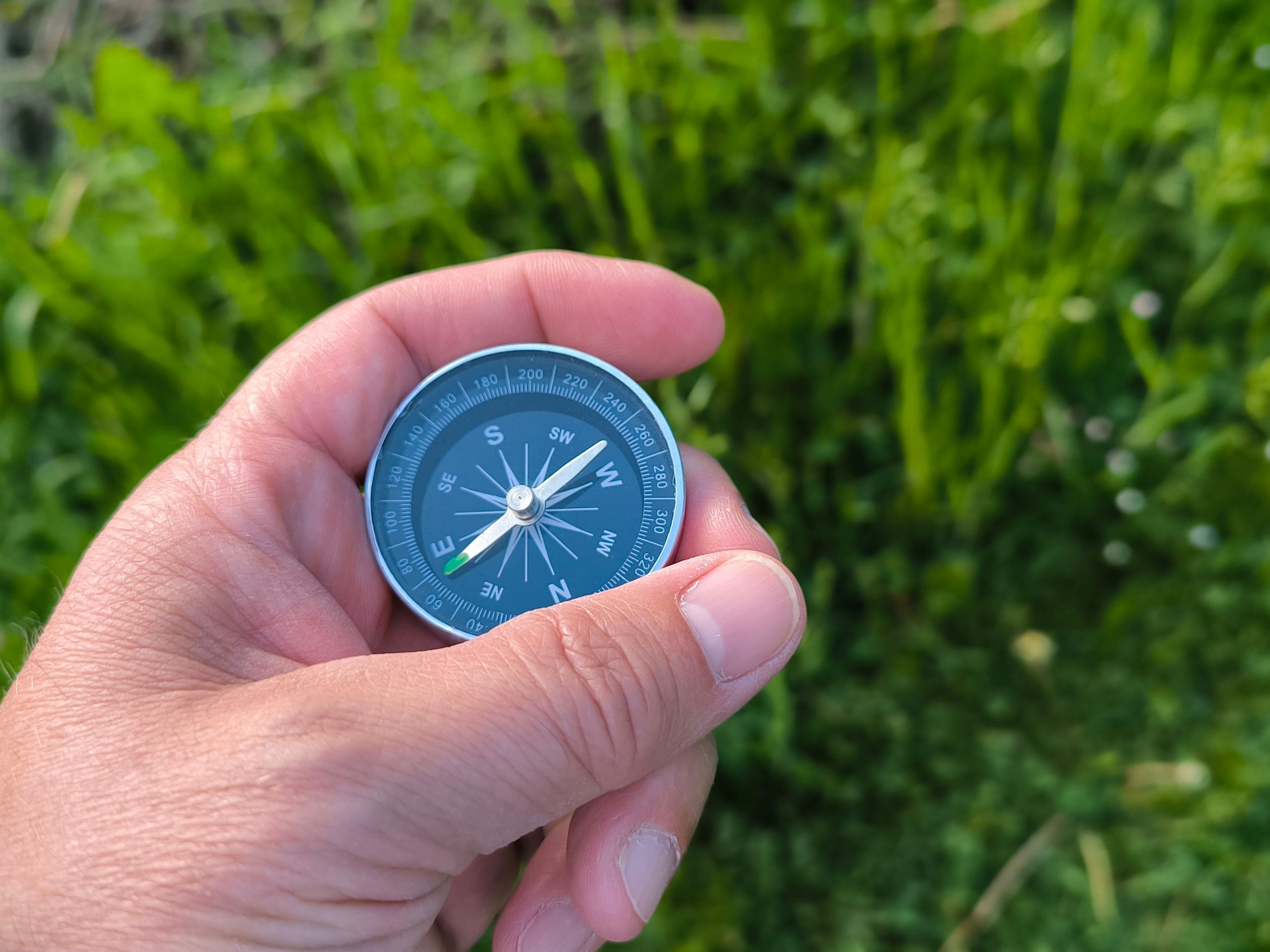 Close-up of a hand holding a compass with a green field in the background. Gros plan d'une main tenant une boussole avec un champ vert en arrière-plan.