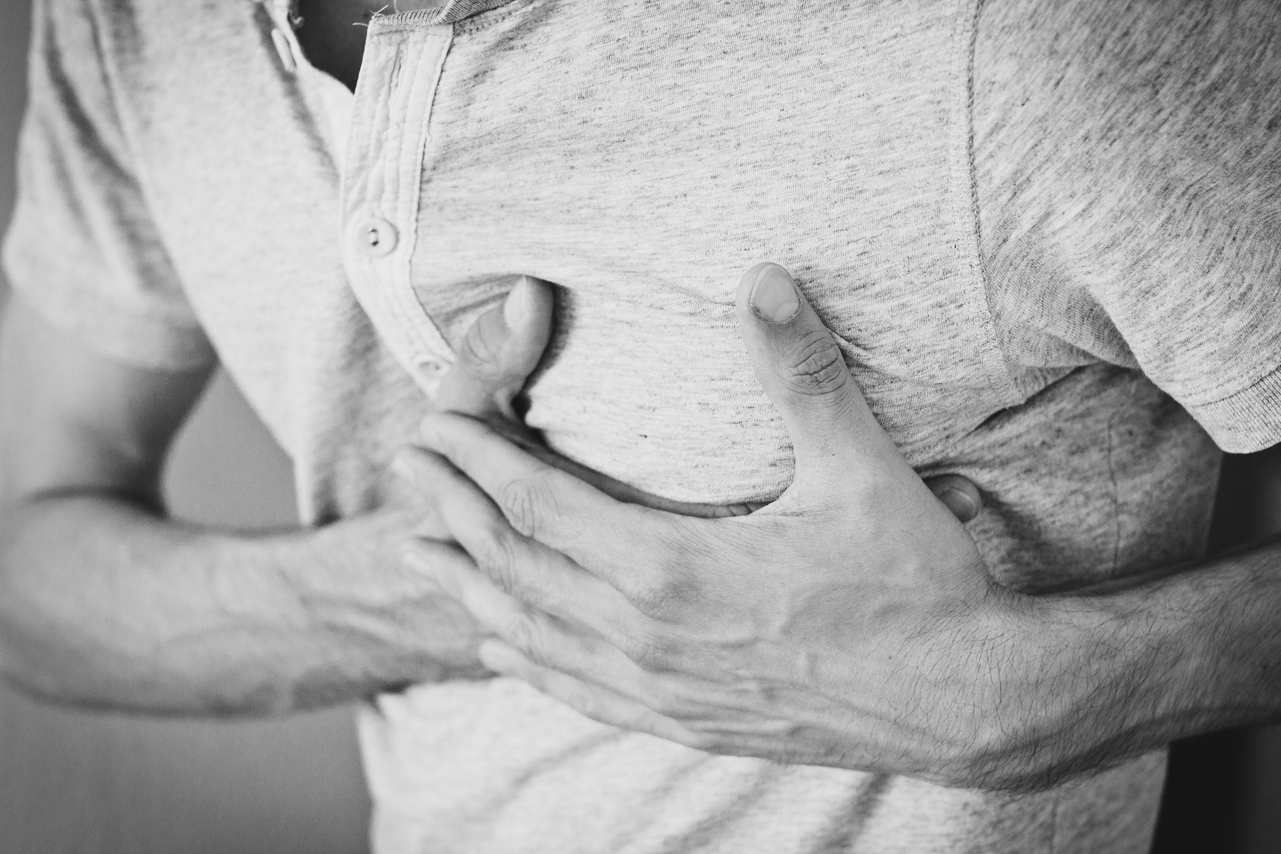Black and white photo of a man clutching his chest, indicating discomfort or pain. Photo en noir et blanc d'un homme se tenant la poitrine, indiquant un malaise ou une douleur.